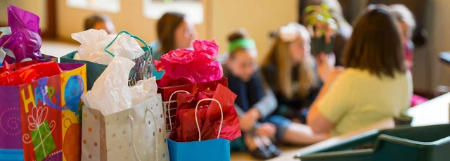 Birthday packages in the foreground with children at a birthday party in the background. 